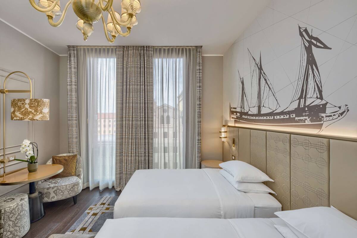 Une chambre d’hôtel avec deux lits, un lustre, une grande fenêtre avec des rideaux légers, une œuvre d’art représentant un voilier accrochée au mur, et une petite table ronde avec une lampe.