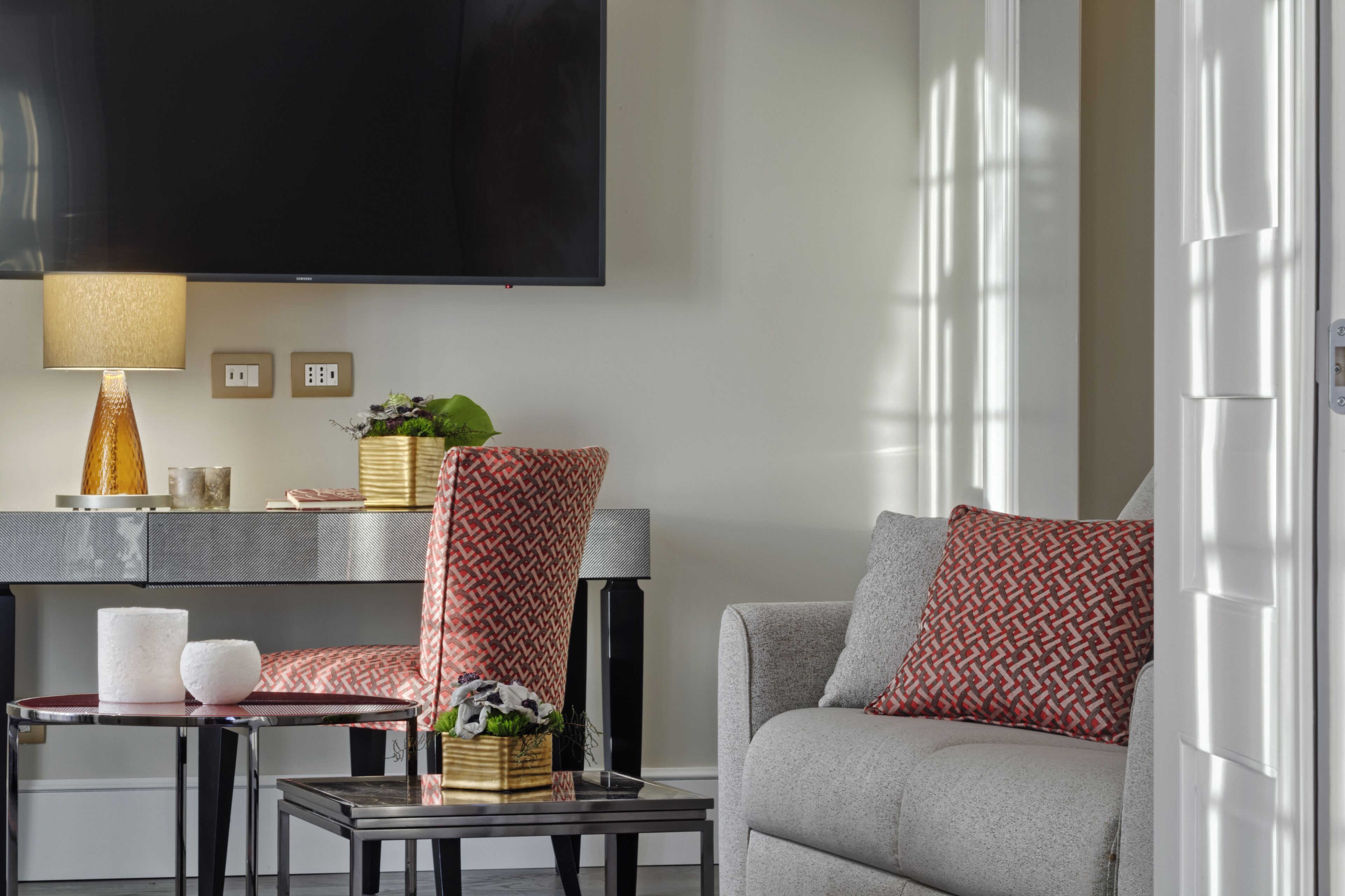 A modern living room with a sofa, armchair, and coffee table. A flat-screen TV is mounted on the wall, and there is a lamp on a side table.