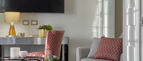 A modern living room with a sofa, armchair, and coffee table. A flat-screen TV is mounted on the wall, and there is a lamp on a side table.
