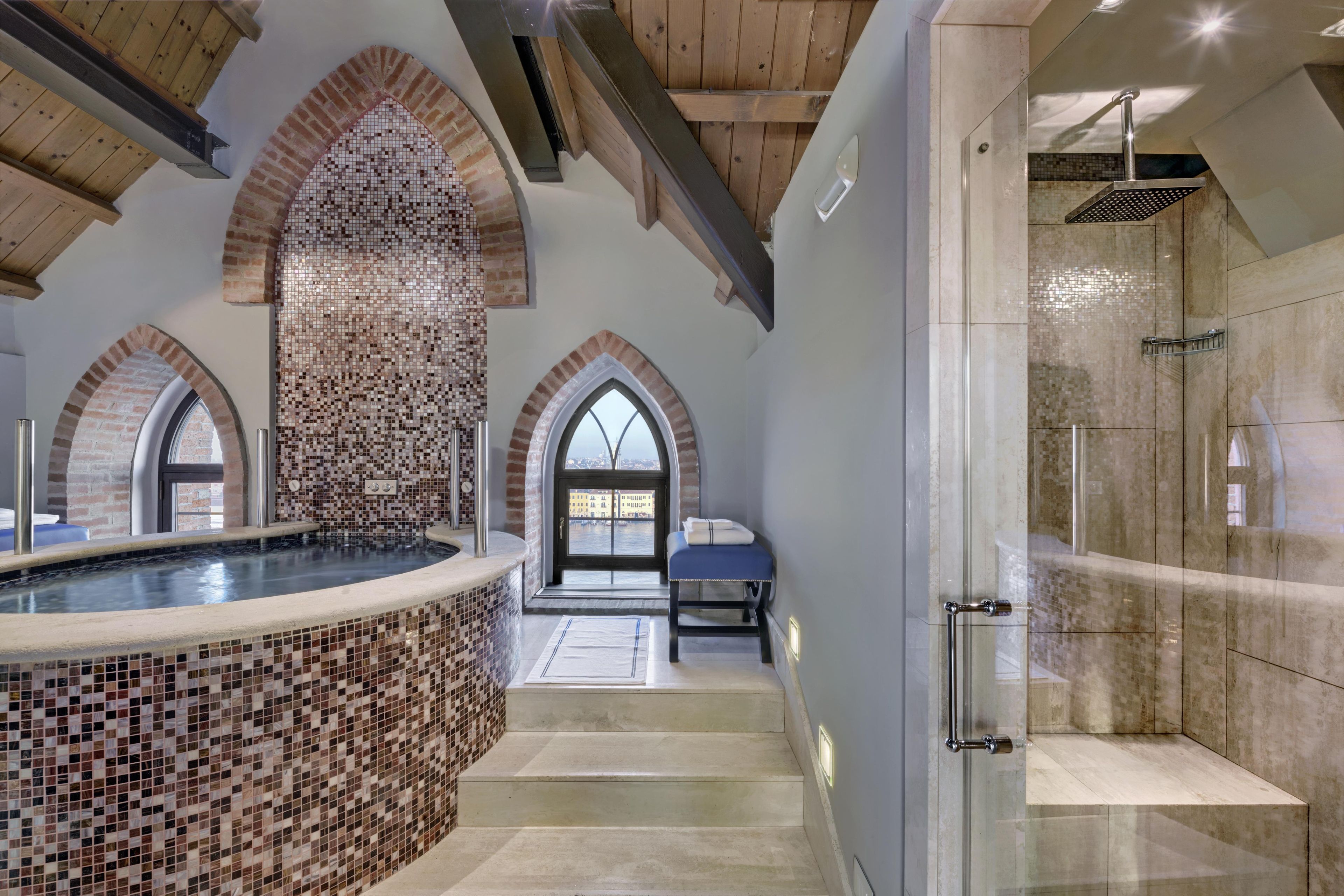 Un espace spa comprenant un bain à remous orné de mosaïques, une douche en verre et une vue sur le paysage urbain, visible à travers une grande fenêtre en arc de cercle.