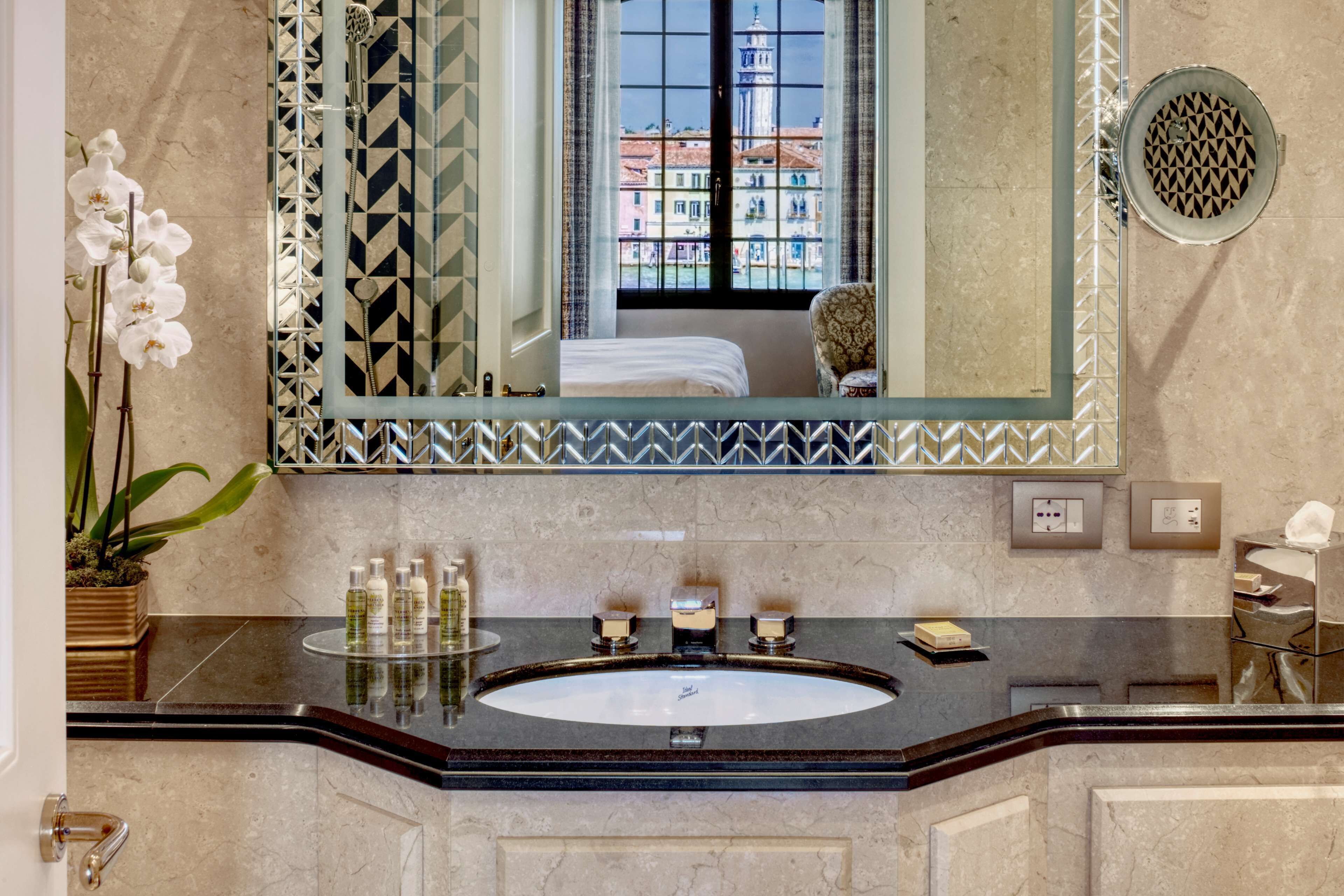 A bathroom with a large mirror, a sink with toiletries, a potted orchid, and a view of buildings through the window.