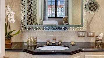 A bathroom with a large mirror, a sink with toiletries, a potted orchid, and a view of buildings through the window.
