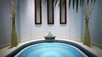 Una piscina con bordo piastrellato, una ciotola di legno con asciugamani arrotolati e due vasi alti con piante secche davanti a una parete con tre specchi.