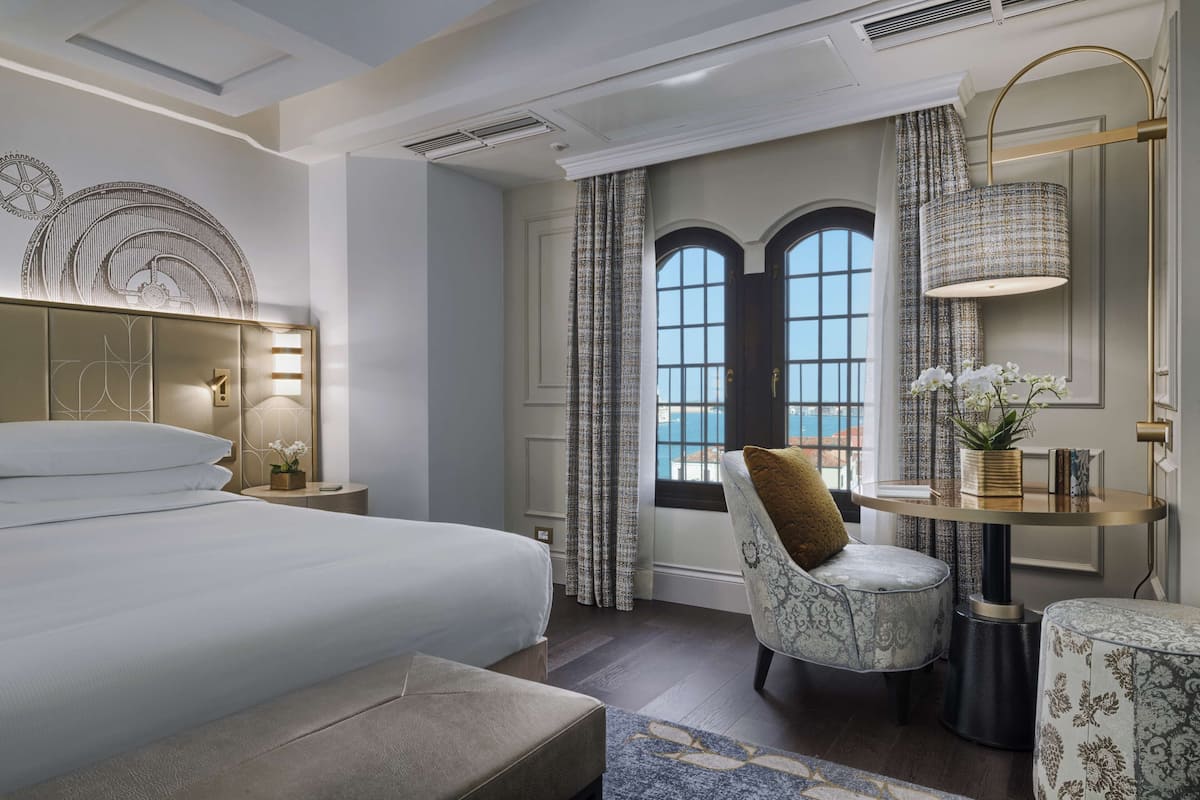 Une chambre d’hôtel avec un grand lit, une table ronde avec une lampe, une chaise et une vue sur l’extérieur par une fenêtre munie de rideaux.