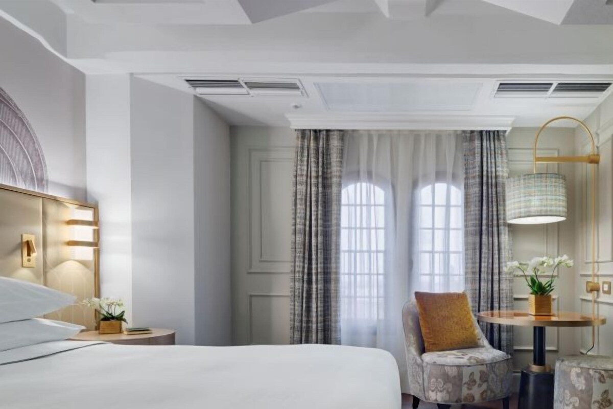 Una habitación de hotel con cama, una silla, una mesa con lámpara y un ventanal con cortinas.
