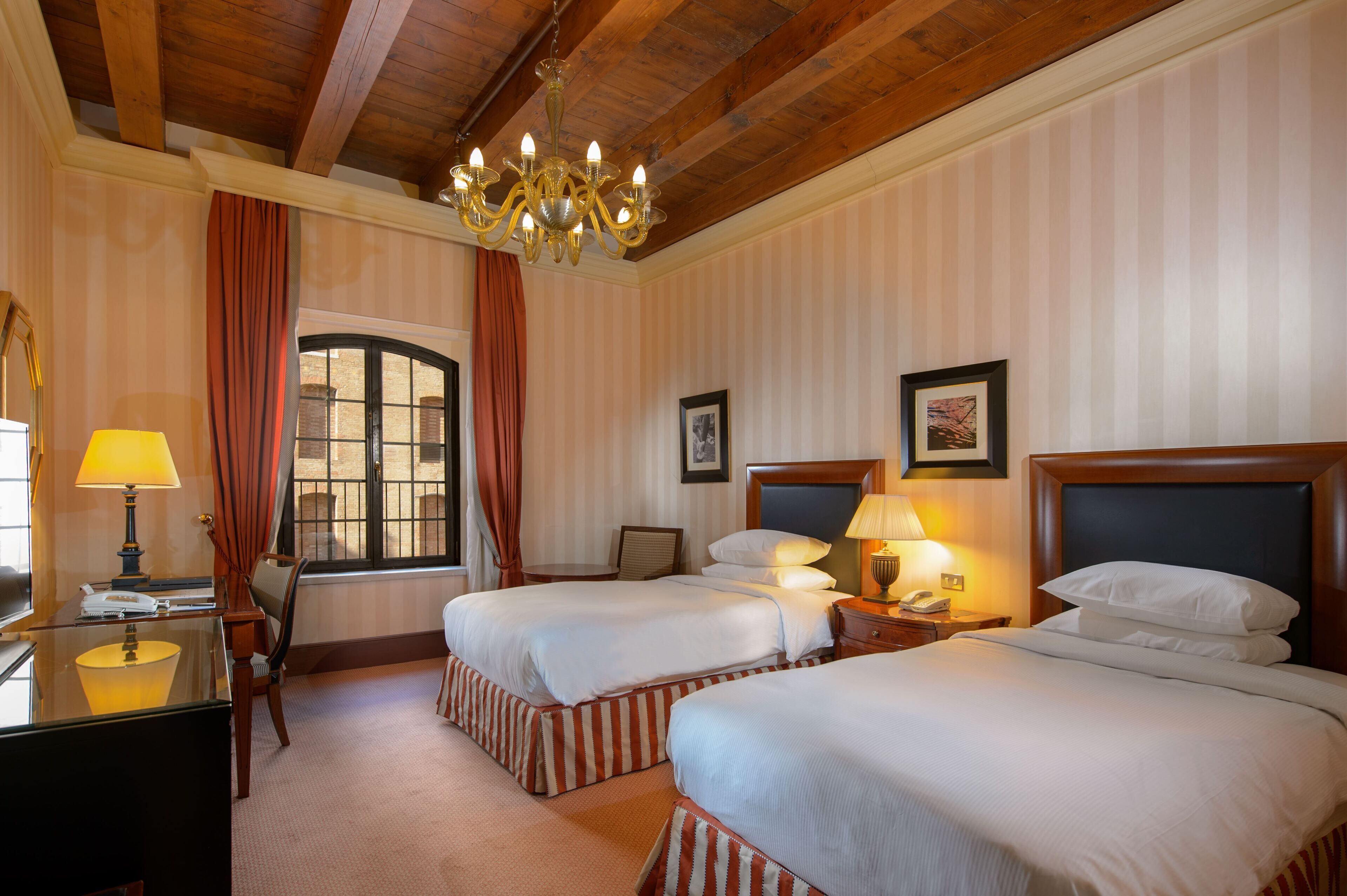 A hotel room with two beds, a chandelier, a desk with a lamp, and a window with curtains.