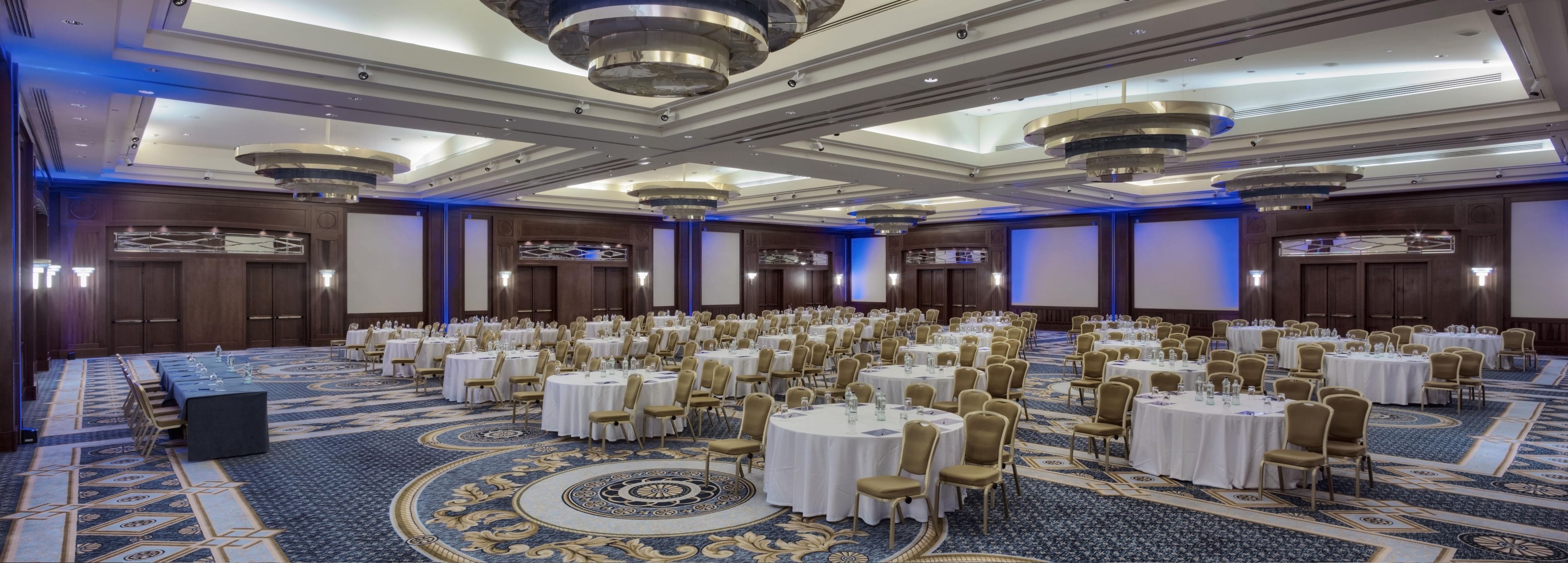 A large conference room with multiple round tables, chairs, and a carpeted floor.
