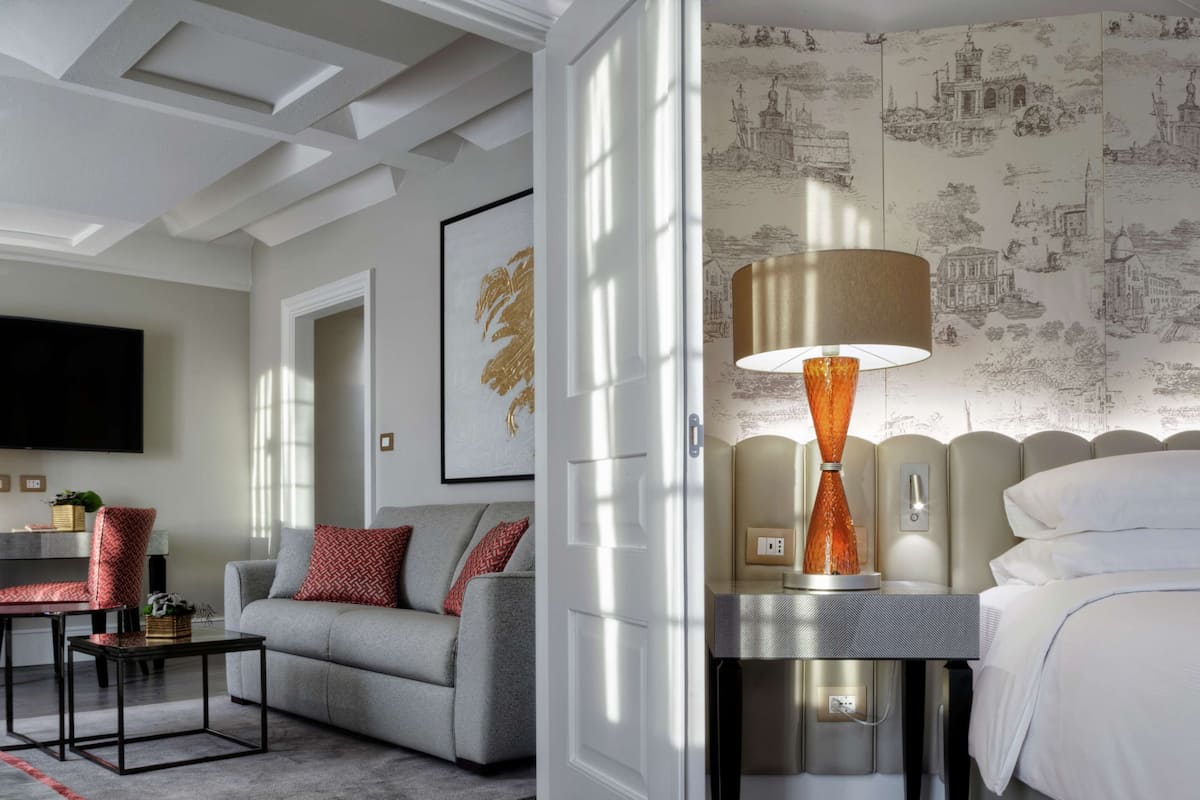 Une chambre d’hôtel moderne avec un canapé gris, un téléviseur à écran plat, une table de chevet et une lampe dotée d’un abat-jour texturé.