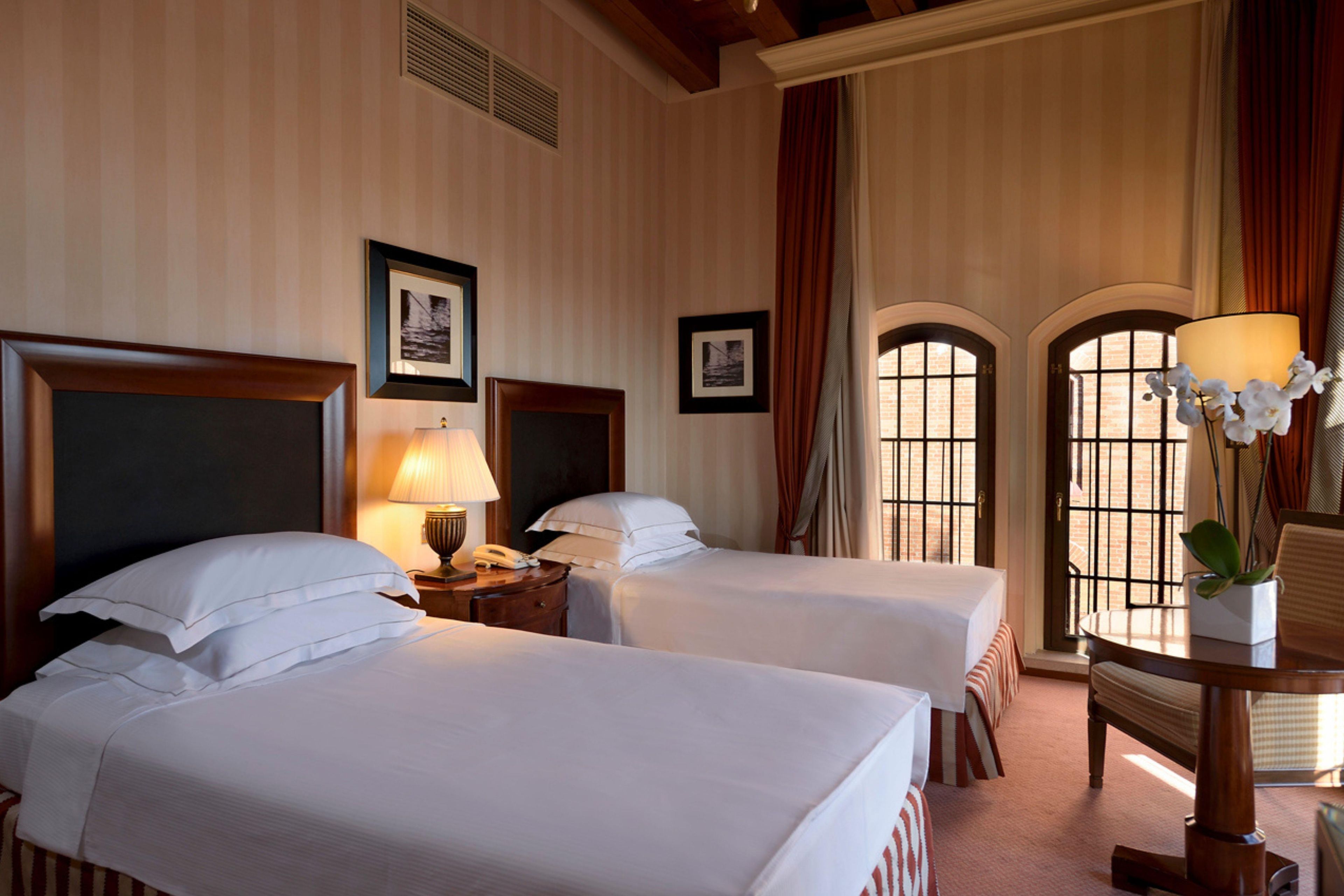 A hotel room with two beds, a nightstand with a lamp, a framed picture, and a window with curtains.