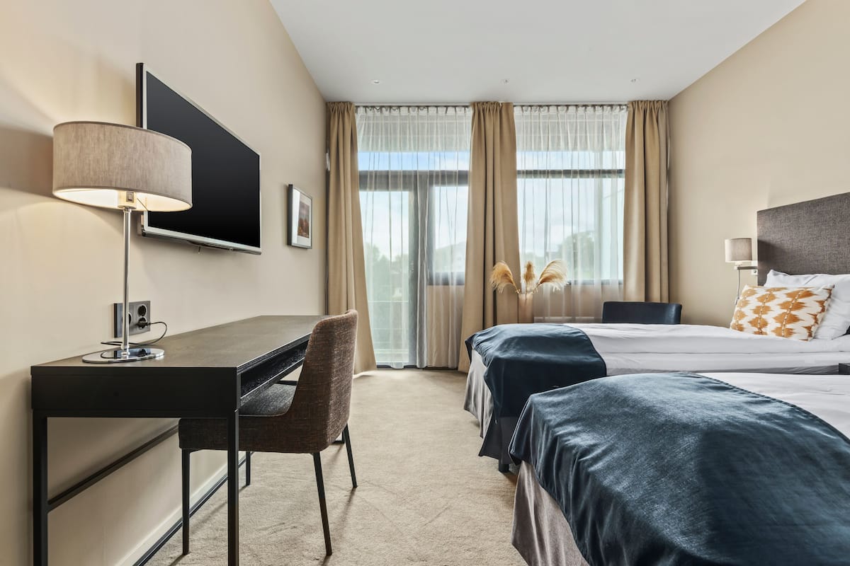 A hotel room with a desk, two beds, a TV, and a large window with curtains.