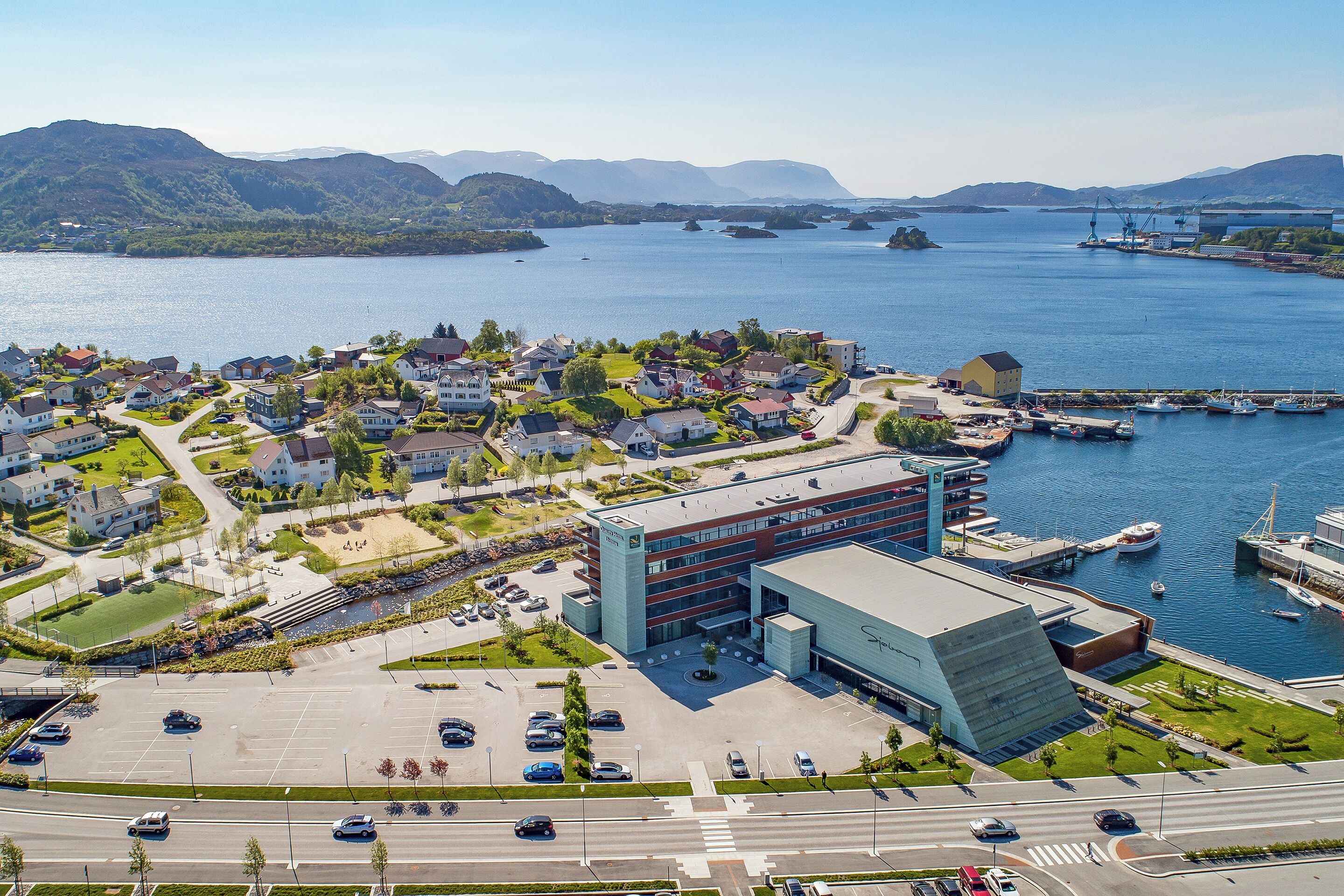 Vista aérea de un pueblo costero con un puerto deportivo, edificios residenciales y un edificio moderno grande con un estacionamiento.