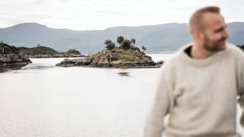 Une personne se promène au bord d’un lac paisible, bordé d’îlots rocheux et surplombant des montagnes lointaines.