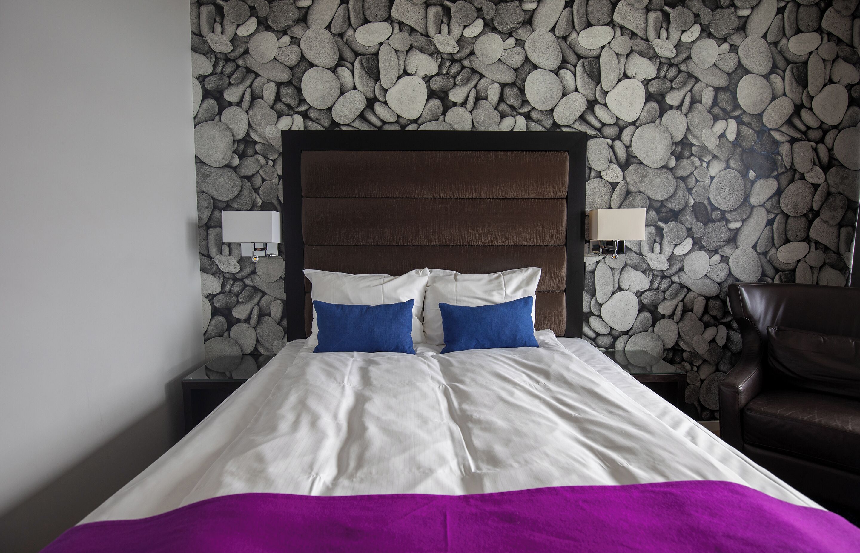 A bed with a headboard, two pillows, a purple blanket, and a wall with a stone pattern.