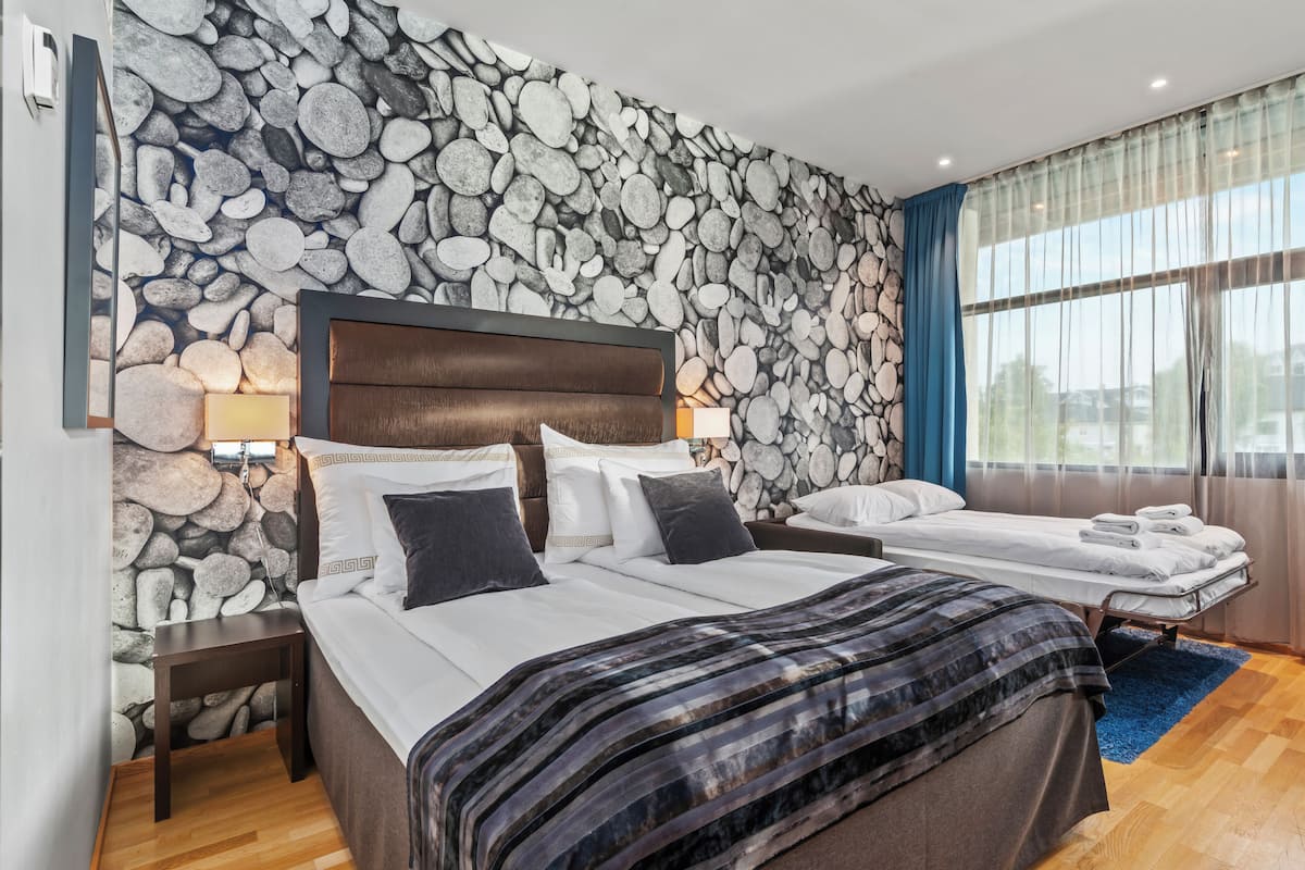 A bedroom with a stone wall, two beds, wooden headboard, bedside tables, lamps, and a window with curtains.