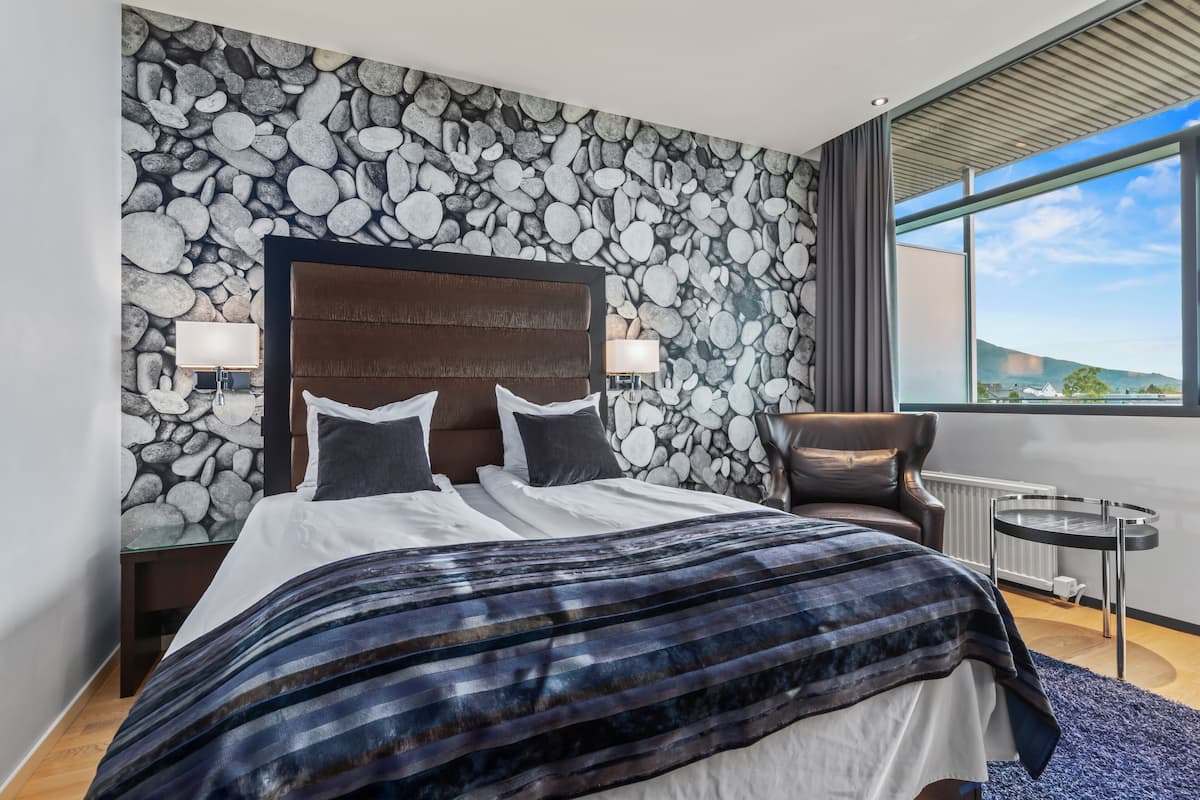 A modern bedroom with a stone wall, a large bed with a striped blanket, a leather chair, and a view of the outdoors through a large window.