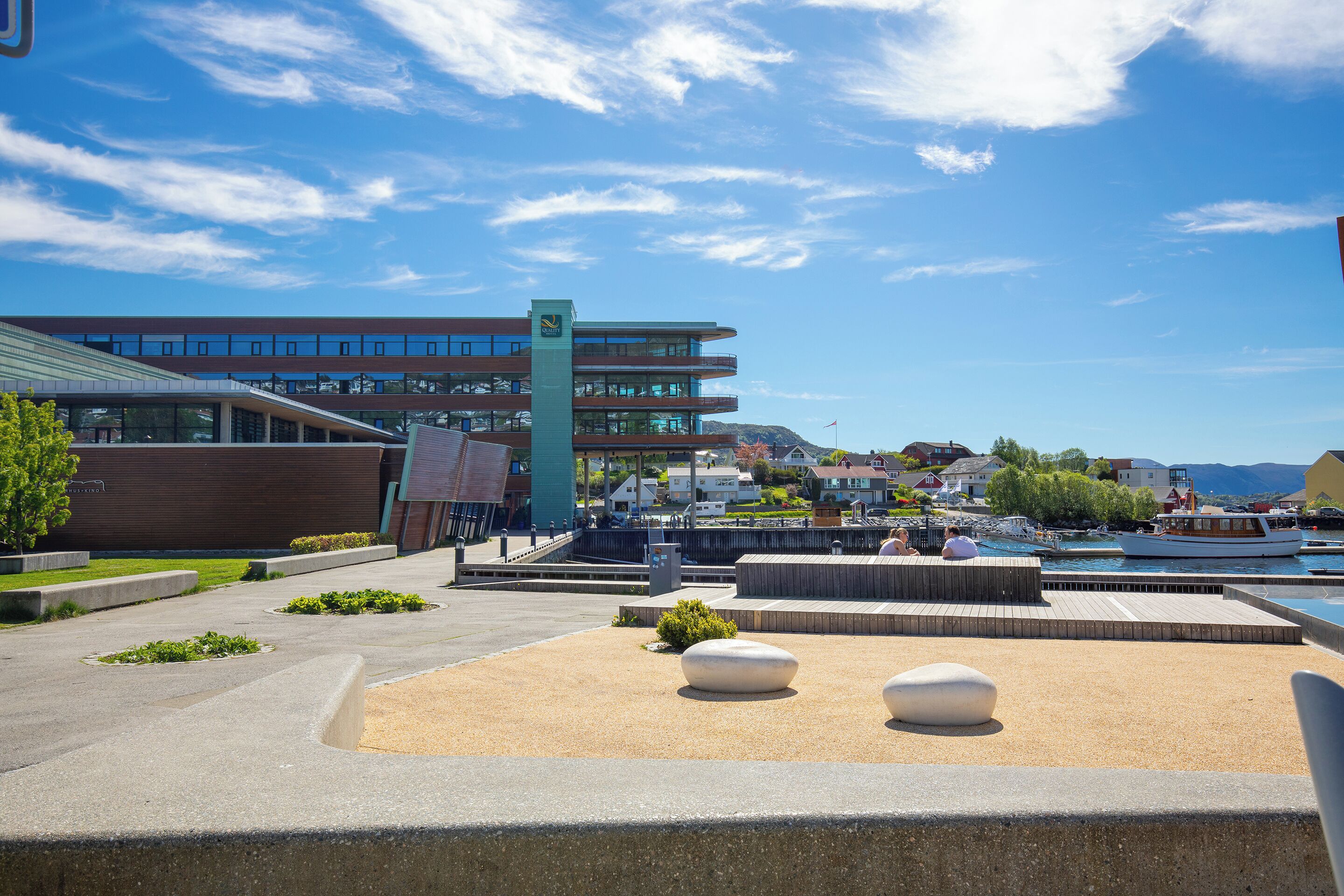 A modern building with multiple floors, a waterfront view, and a public square with seating areas.