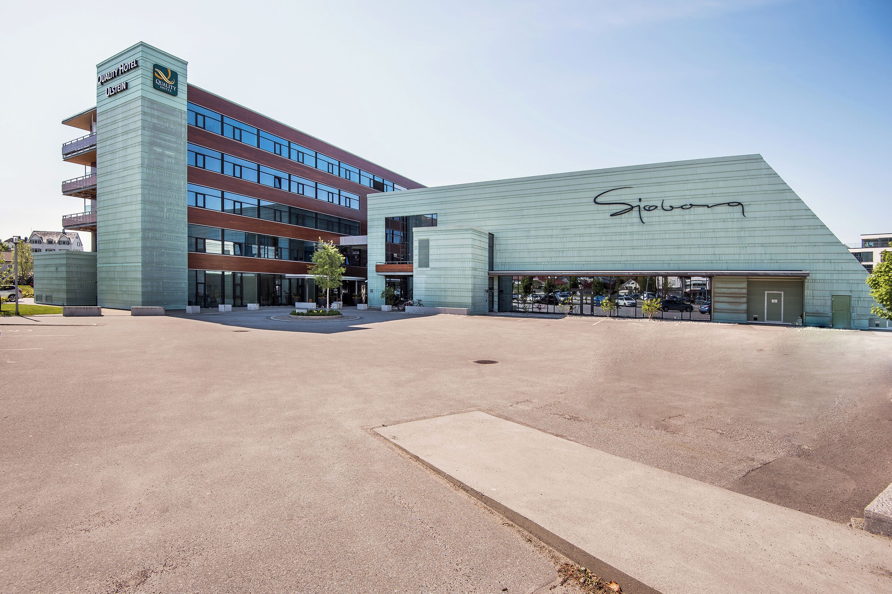A modern hotel with a prominent 'Quality Hotel' sign, a large 'Sjoborg' sign, and a spacious outdoor area with a clear sky.