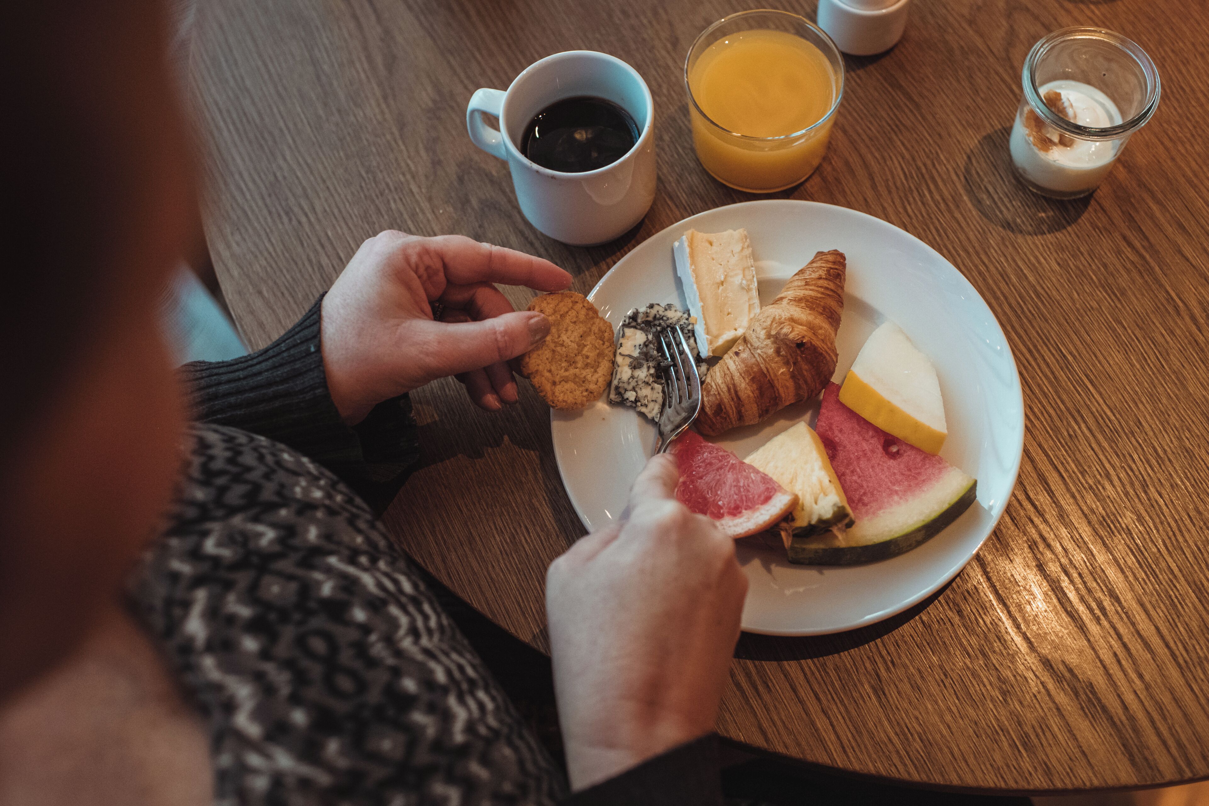 A breakfast plate with croissant, cheese, and fruit, a cup of coffee, and a glass of orange juice on a wooden table.