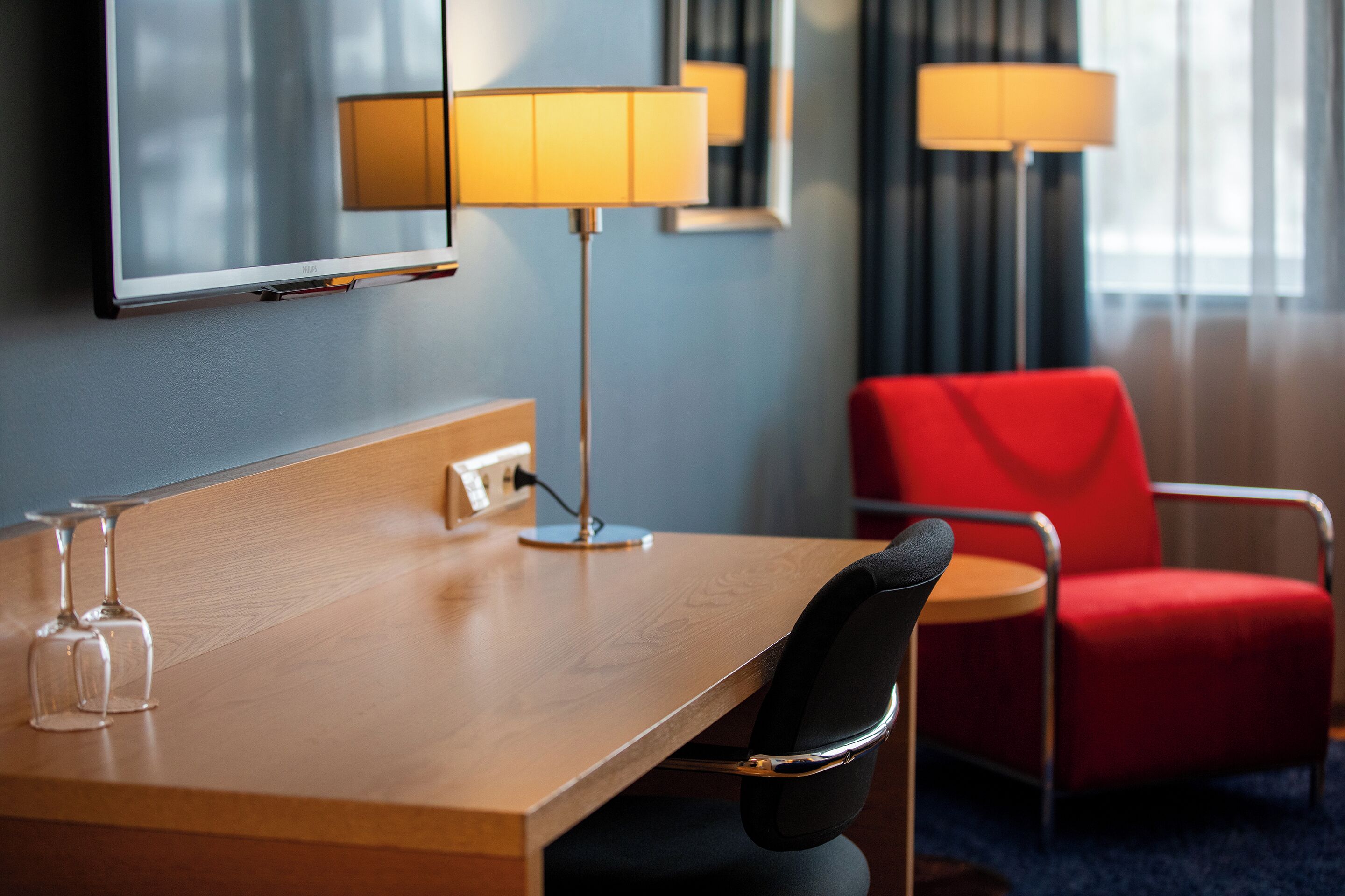 A hotel room with a wooden desk, two lamps, a red chair, and a television.