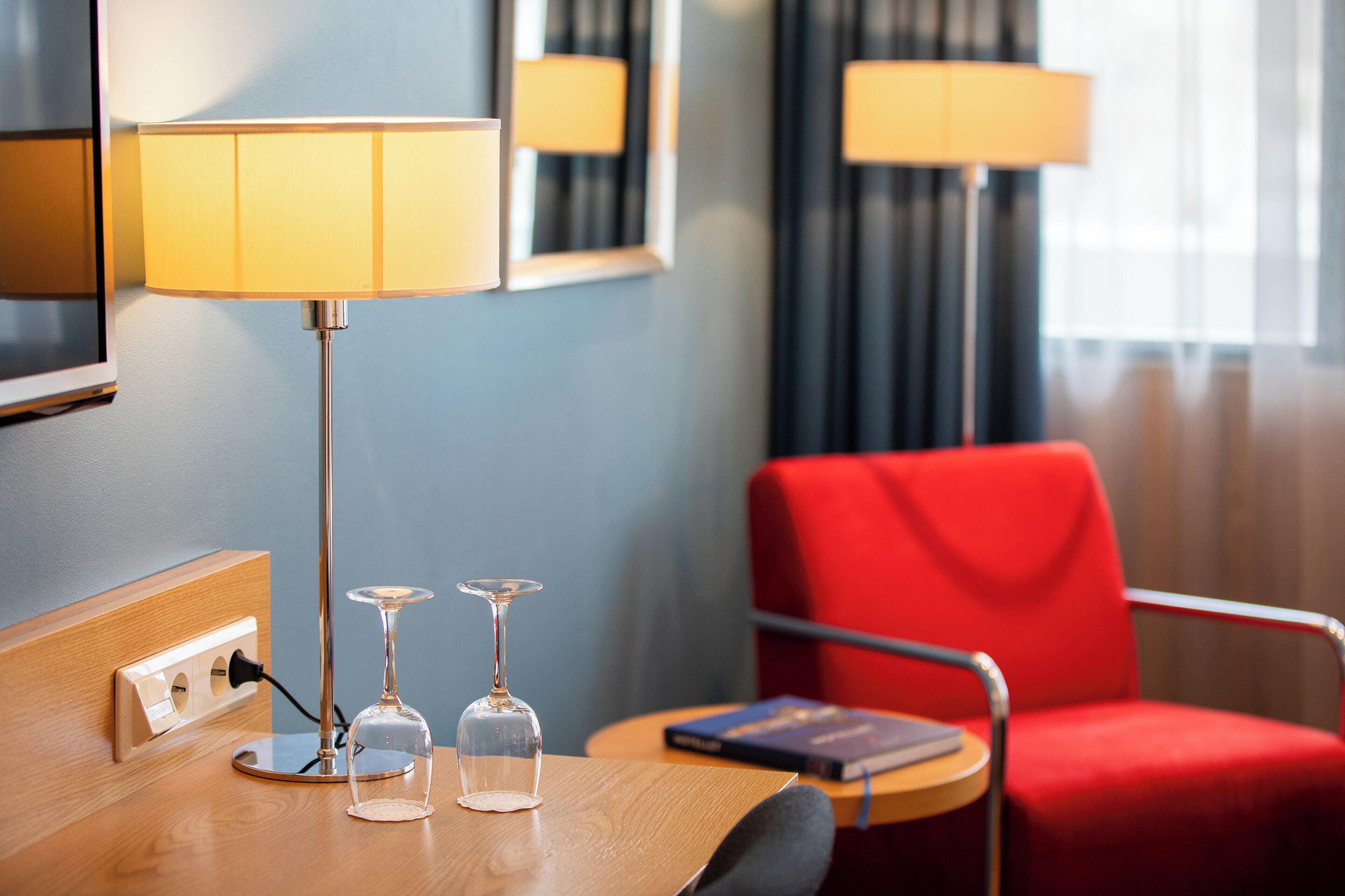 Et hotelværelse med et træskrivebord, to vinglas, en lampe, en rød stol og en bog.