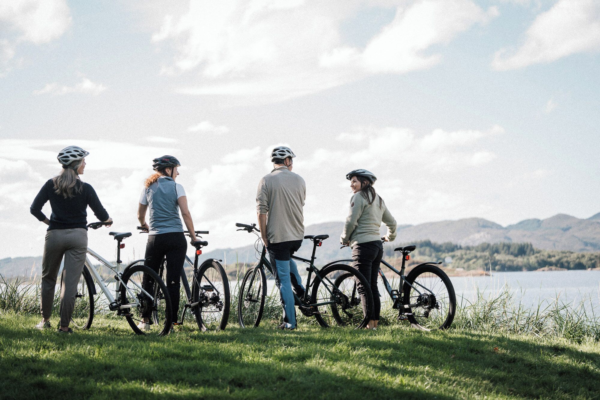 Cuatro personas de pie sobre césped, cerca de bicicletas, con vistas a un cuerpo de agua y montañas.