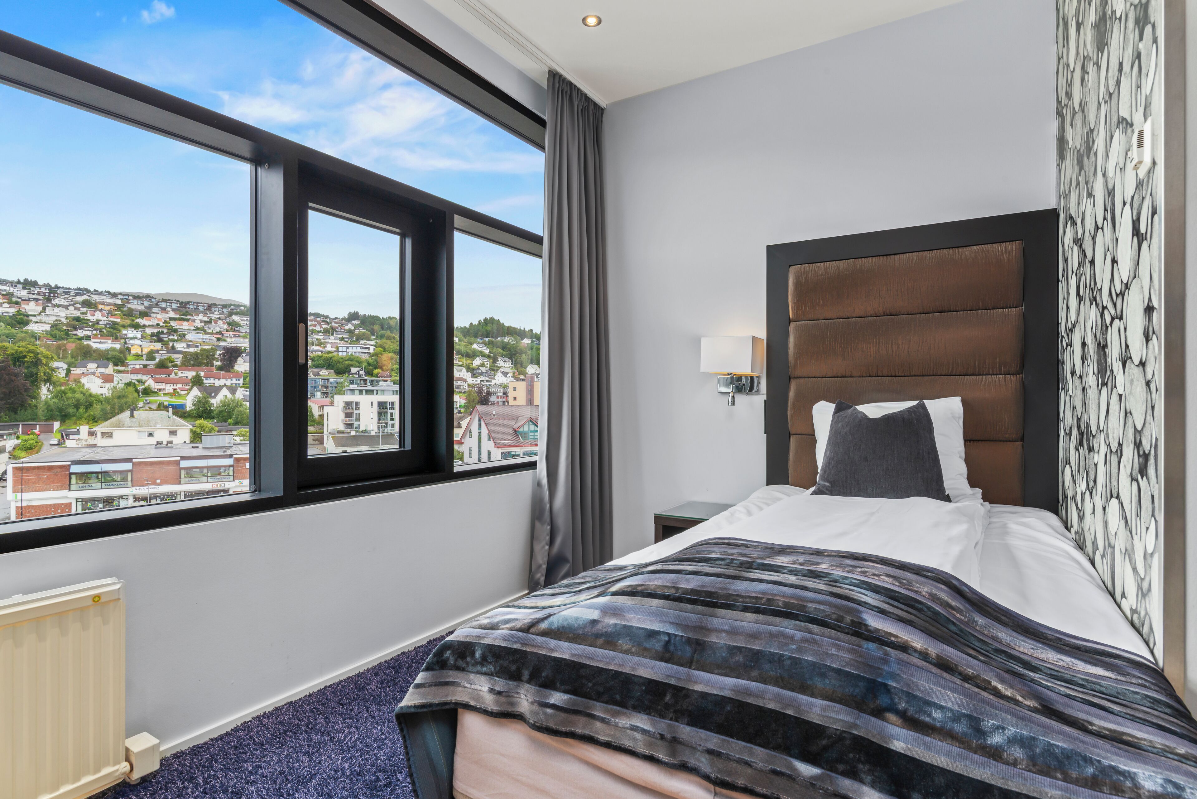 A hotel room with a large window, a bed with a striped blanket, a headboard with a patterned wallpaper, and a nightstand with a lamp.