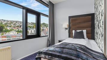 A hotel room with a large window, a bed with a striped blanket, a headboard with a patterned wallpaper, and a nightstand with a lamp.