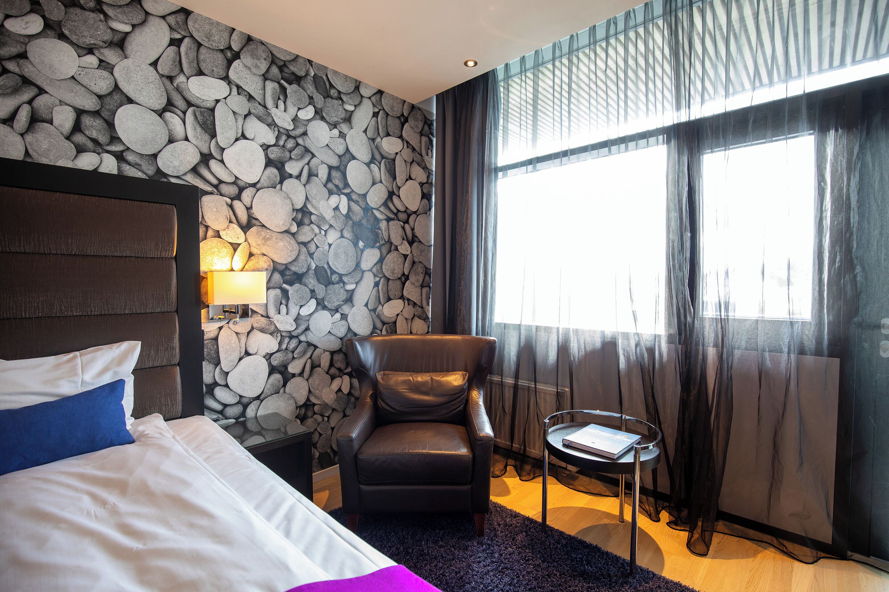 A hotel room with a bed, a chair, a small table, a lamp, a stone wall, and a window with curtains.