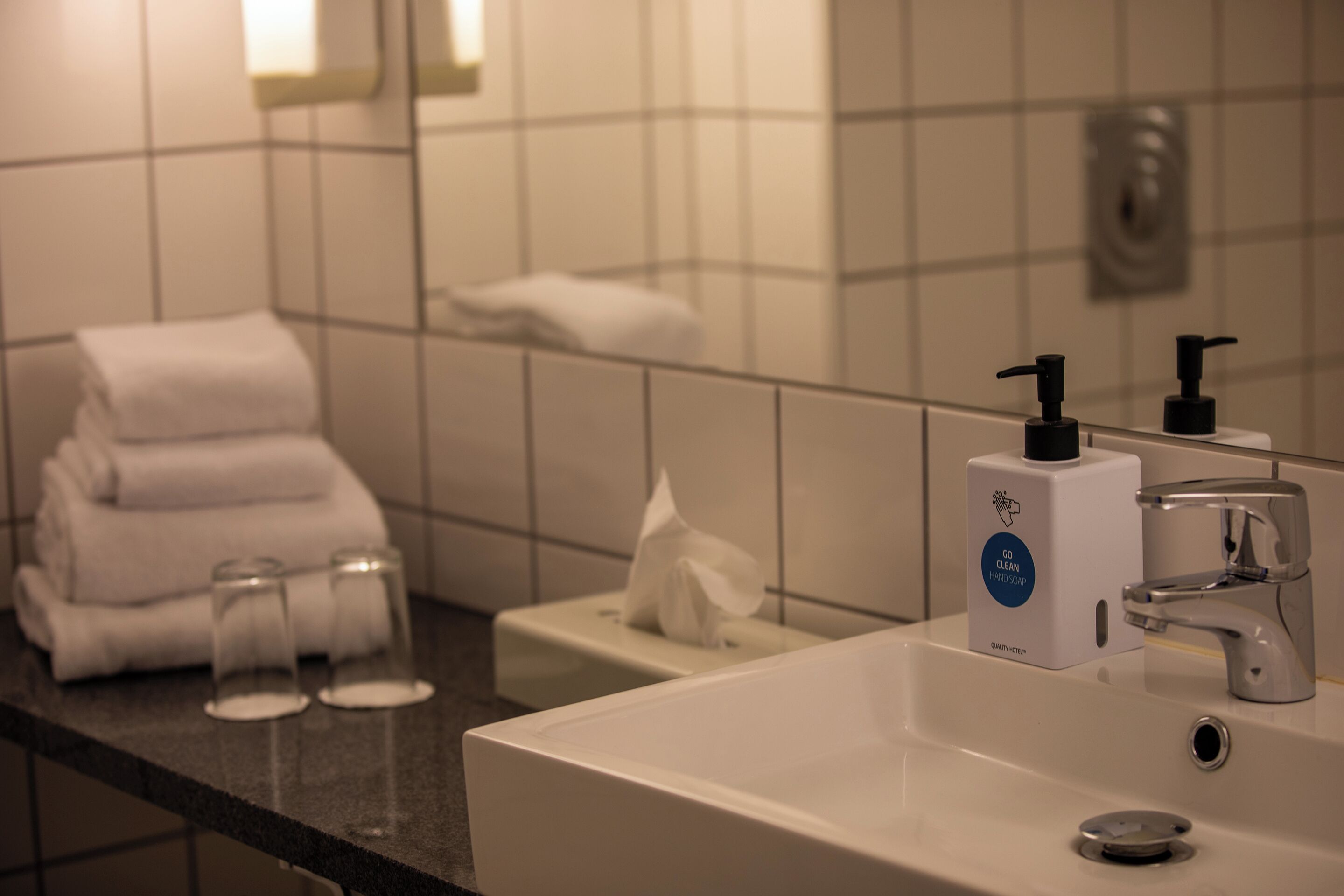 A hotel bathroom with a sink, hand soap dispenser, and a towel stack.