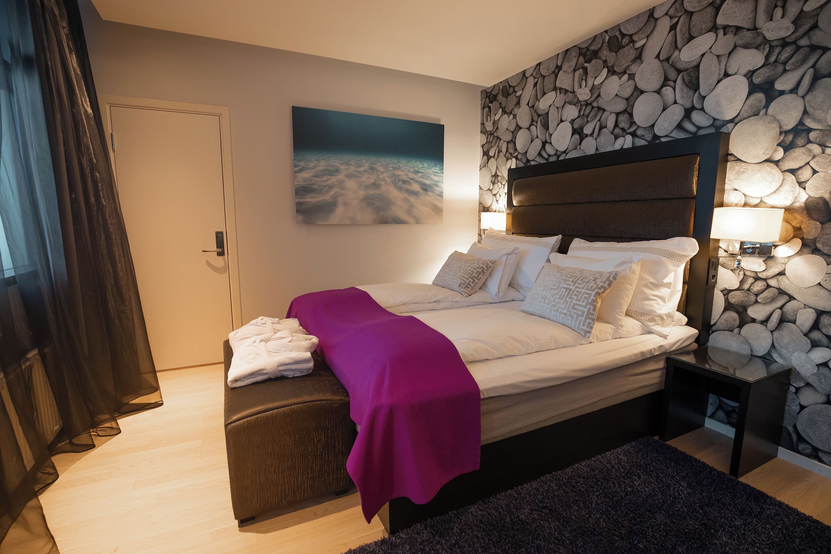 Ein Schlafzimmer mit Bett, Nachttisch, Bank, einer Wand mit Steinmuster und einem Strandbild.