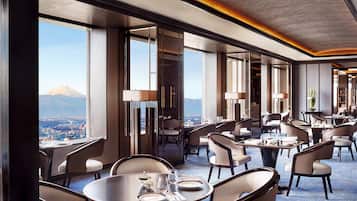 A hotel dining area with tables and chairs, large windows with a mountain view, and a modern interior design.