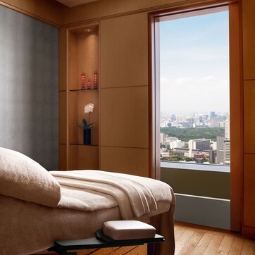 A modern room with a large window, a bed with a beige blanket, a small table with a white object, and a wall-mounted shelf with decorative items.