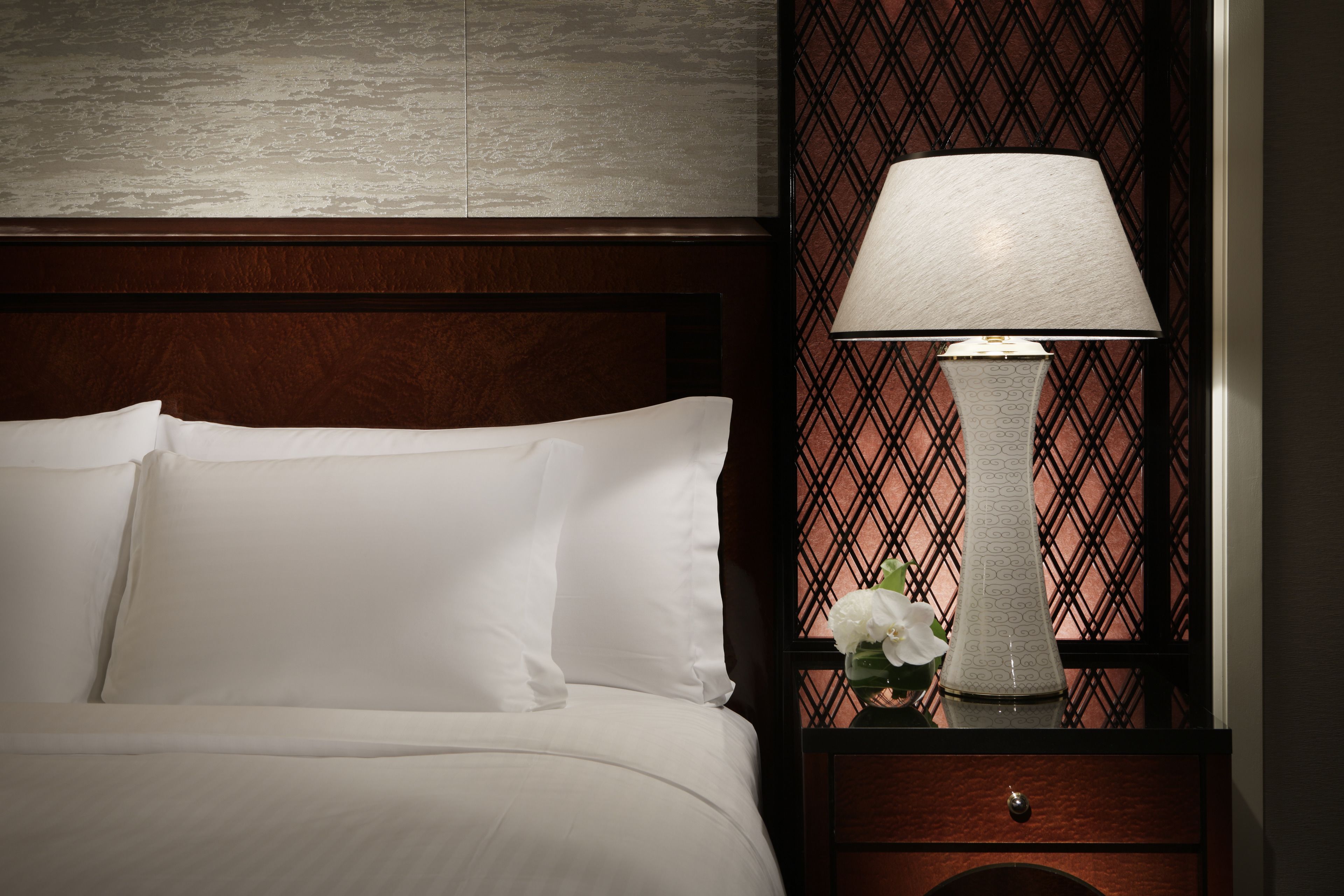 A neatly made bed with white linens, a bedside table with a lamp, and a decorative panel with a geometric pattern.