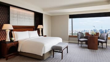 Habitación de hotel con una cama grande, dos mesitas de noche con lámparas, una pequeña mesa con sillas y vistas a un paisaje urbano.