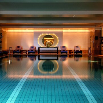 A modern indoor swimming pool with a central heart-shaped light installation, lounge chairs, and a reception desk.
