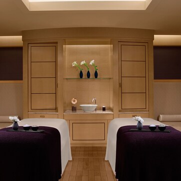 A spa room with two treatment beds, a central sink, and a shelf with decorative items.