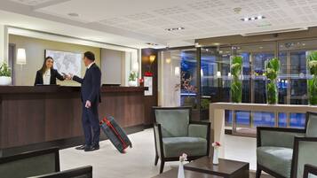 Een hotel receptie met een balie, stoelen, een klein tafeltje met een vaas, en uitzicht op buiten door grote ramen.