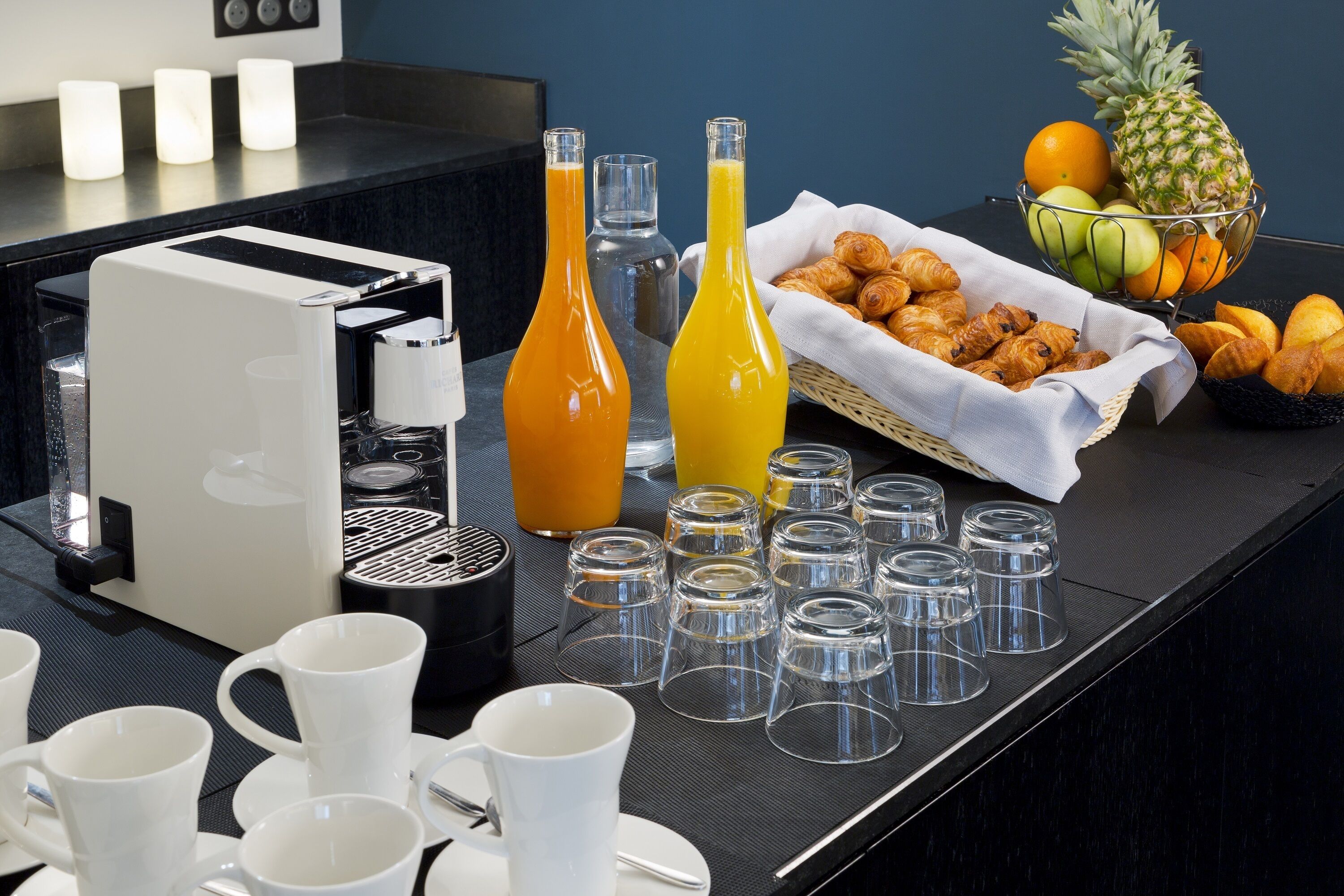 Un plateau de petit-déjeuner comprenant une machine à café, des bouteilles de jus, des croissants et des fruits.