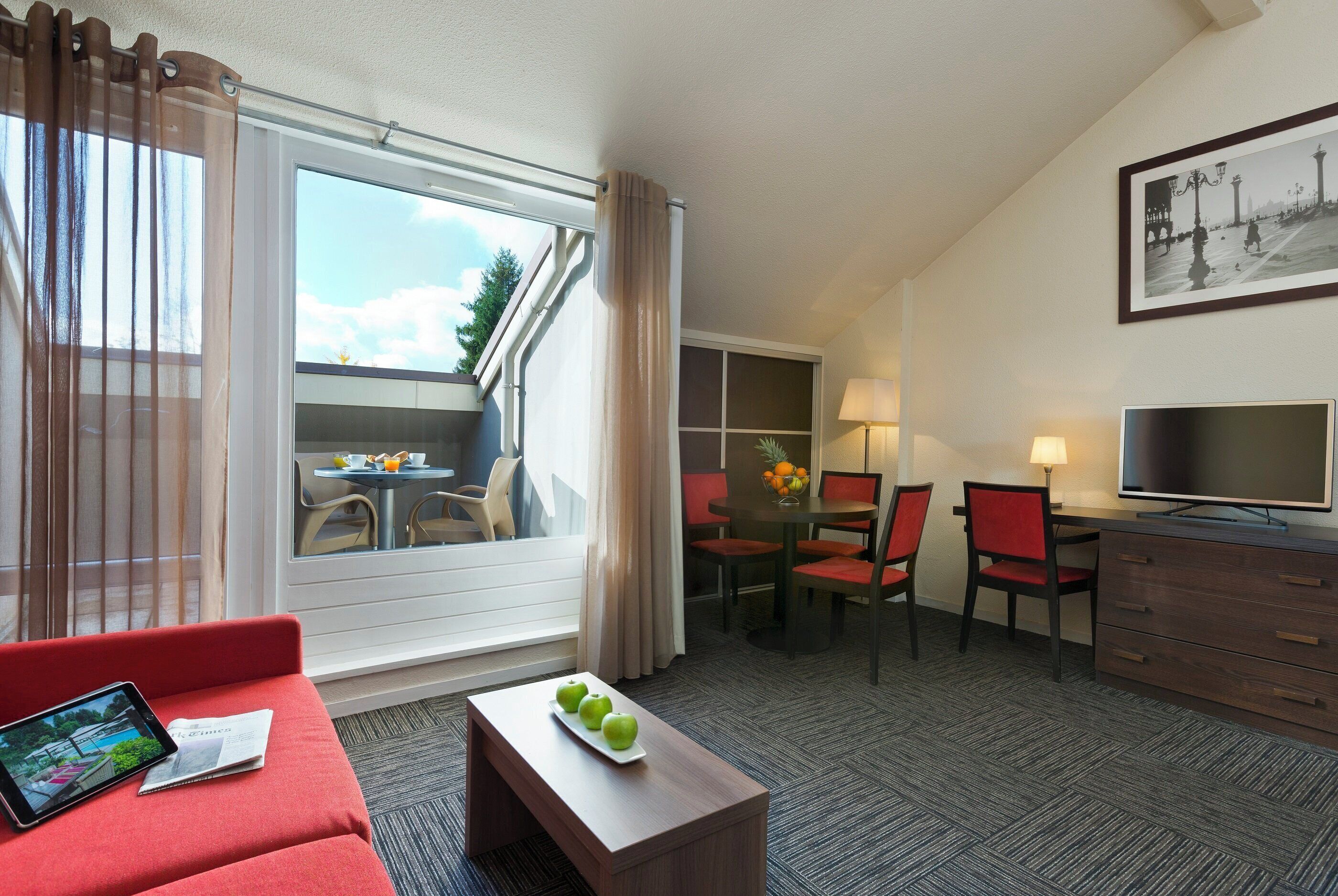 Quarto de hotel com um sofá vermelho, uma mesa pequena com maçãs, uma televisão, área de refeições com cadeiras e uma janela grande com cortinas.