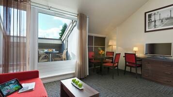 Quarto de hotel com um sofá vermelho, uma mesa pequena com maçãs, uma televisão, área de refeições com cadeiras e uma janela grande com cortinas.
