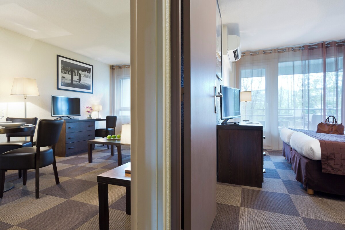 Una habitación de hotel con cama, escritorio, silla, televisión y ventana con cortinas.