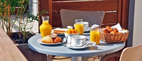 Une table ronde est dressée avec des aliments pour le petit-déjeuner, notamment des croissants, du jus de fruits et un panier de viennoiseries. L’ensemble est placé devant un décor en bois, avec une plante en pot à proximité.