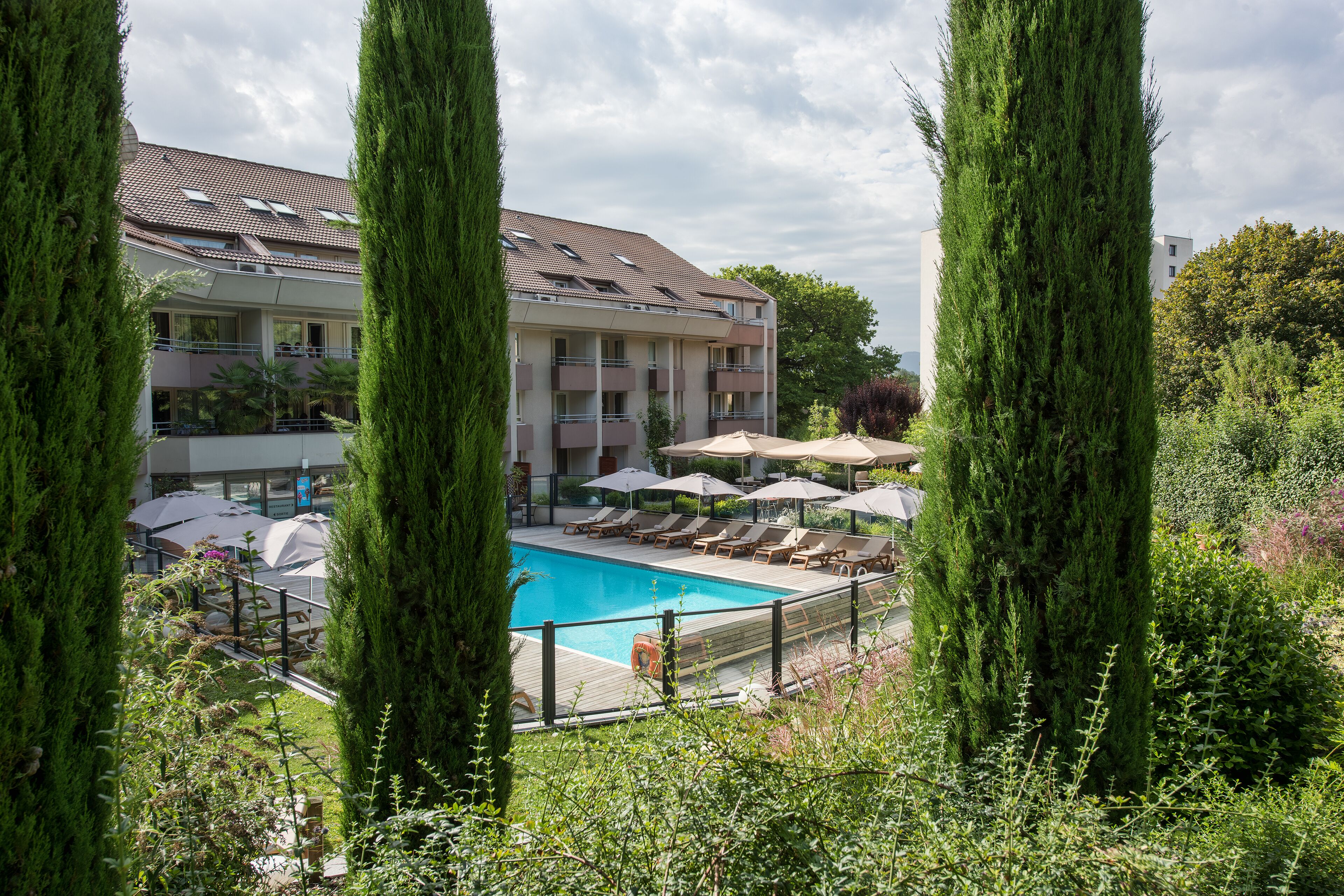 A pool area with lounge chairs and umbrellas, surrounded by tall trees and a building with balconies.