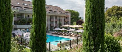 A pool area with lounge chairs and umbrellas, surrounded by tall trees and a building with balconies.