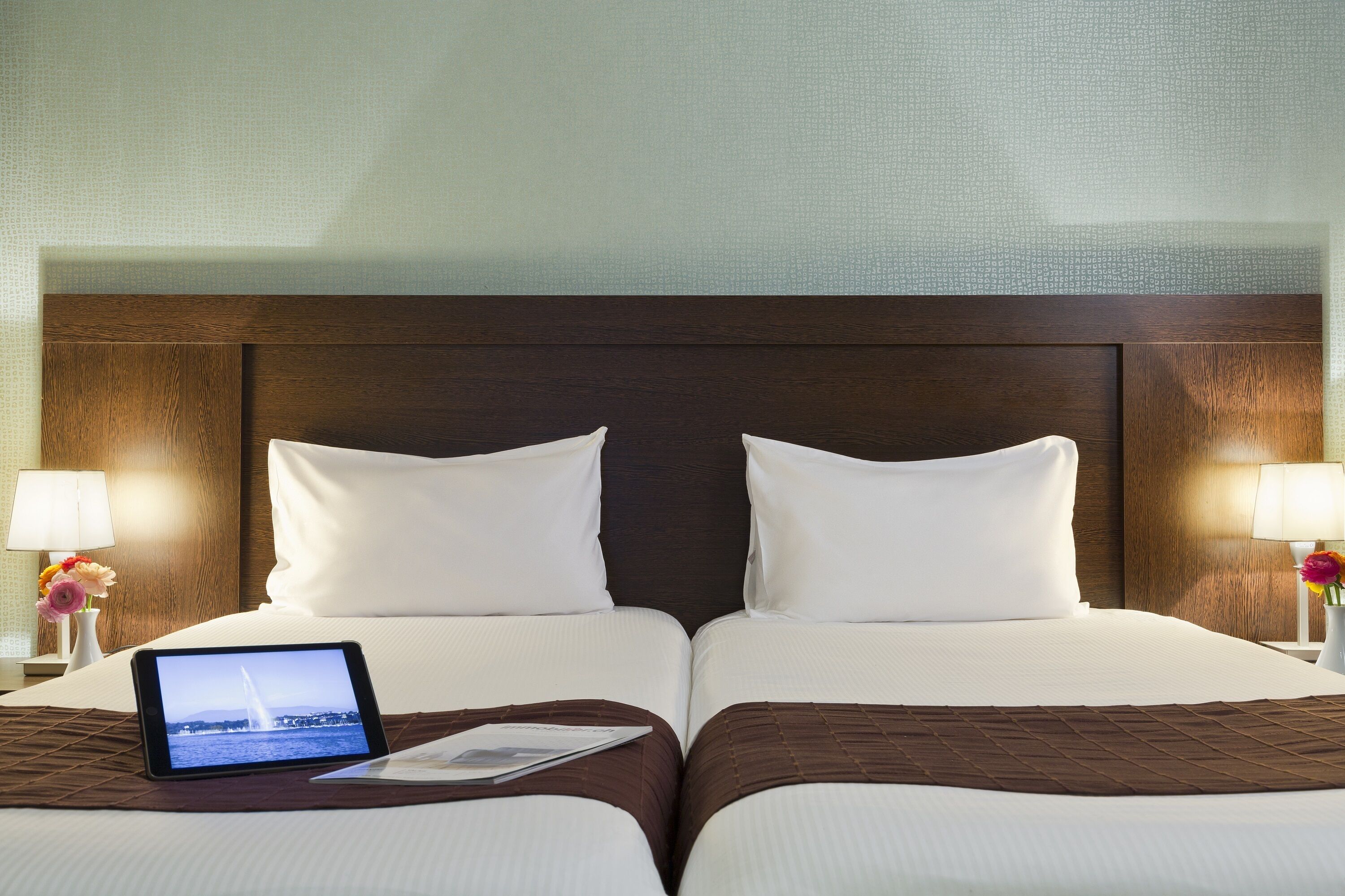 Une chambre d’hôtel avec deux lits, une table de chevet avec une lampe et une tablette affichant une vue panoramique.