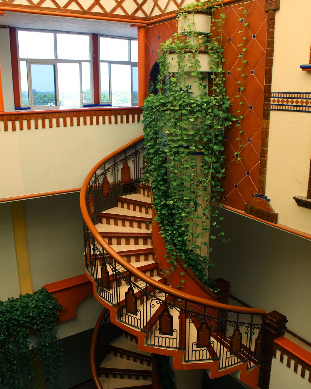 A spiral staircase with a wooden handrail and black balusters, surrounded by walls with a geometric pattern and a large window at the top.