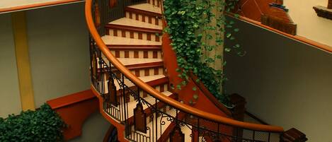 A spiral staircase with a wooden handrail and black balusters, surrounded by walls with a geometric pattern and a large window at the top.