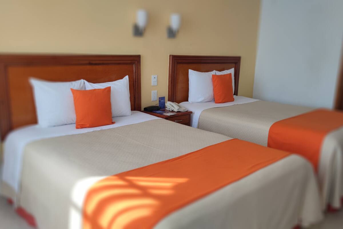 Deux lits doubles avec une literie orange et blanche, une table de chevet avec un téléphone et un livre, et un luminaire fixé au mur.