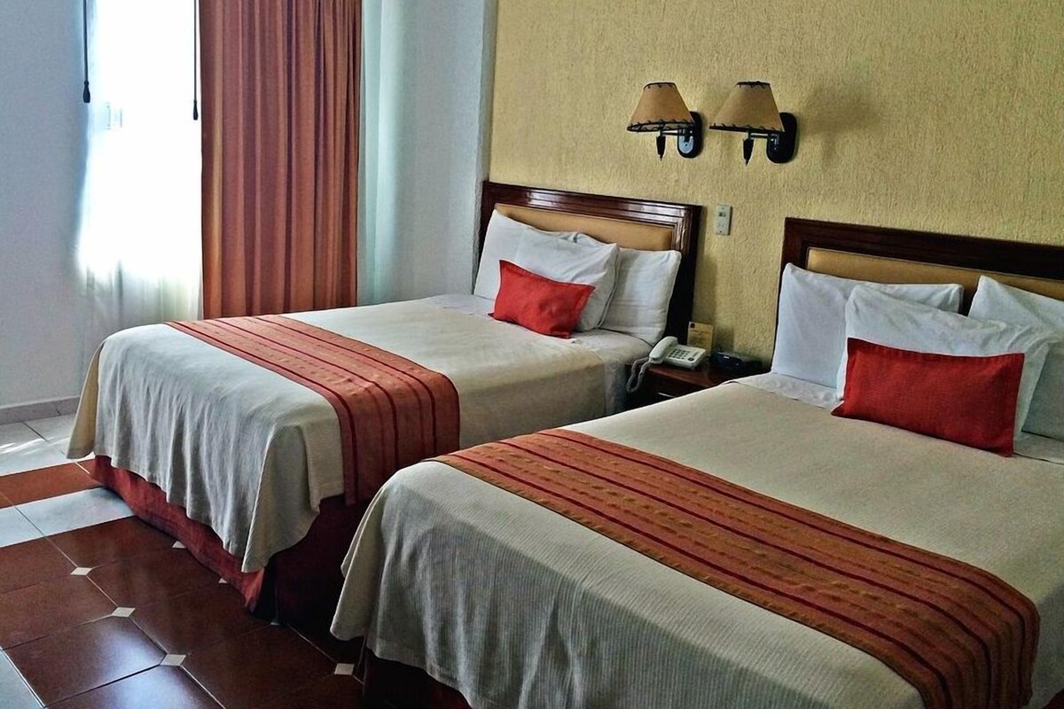 Habitación de hotel con dos camas, cada una con cubrecama rojo y blanco, cabecero de madera y mesita de noche con teléfono.