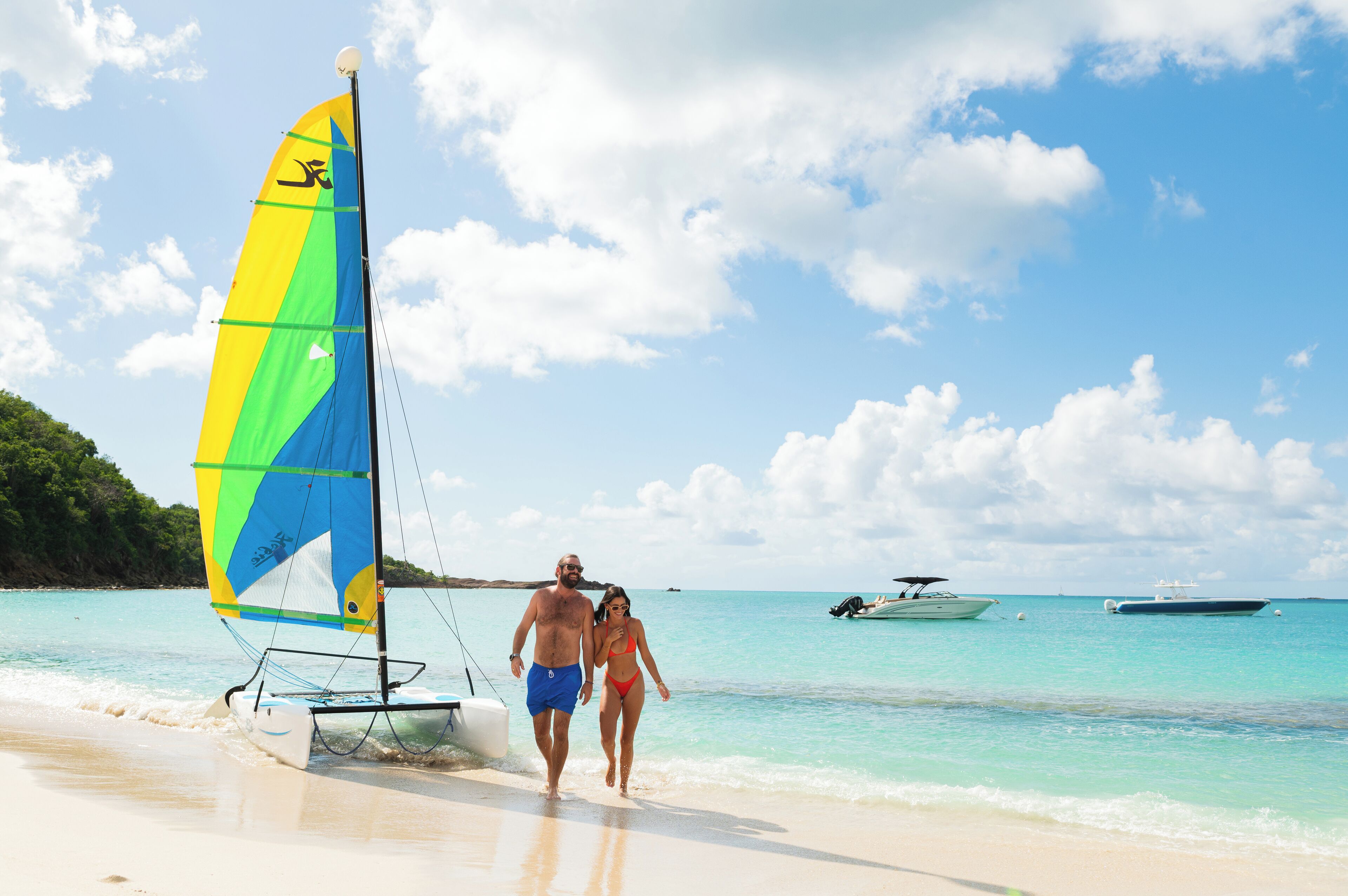 Une plage avec un voilier, un couple qui se promène et des bateaux sur l’eau.